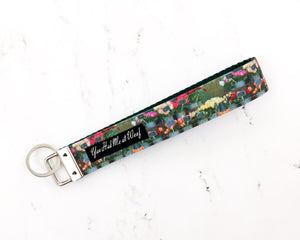 A key fob with dark green webbing and a photographic image of beautiful flowers in galvanized tubs in front of a flower shop, with black You Had Me at Woof label and split O ring to hold keys.